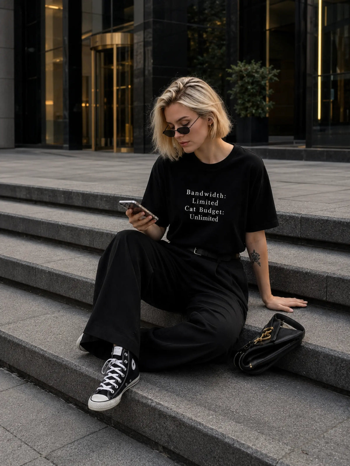 Woman sitting on steps wearing a black t-shirt with text, using a phone.